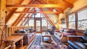 Interior acogedor de una cabaña de madera con techo alto, vigas de madera, una zona de estar central con sofás y una mesa de centro, y ventanas grandes con persianas.