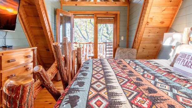 Una cabaña con habitación entre piso, vigas de madera, una cama con ropa de cama estampada, un aparador y un televisor.