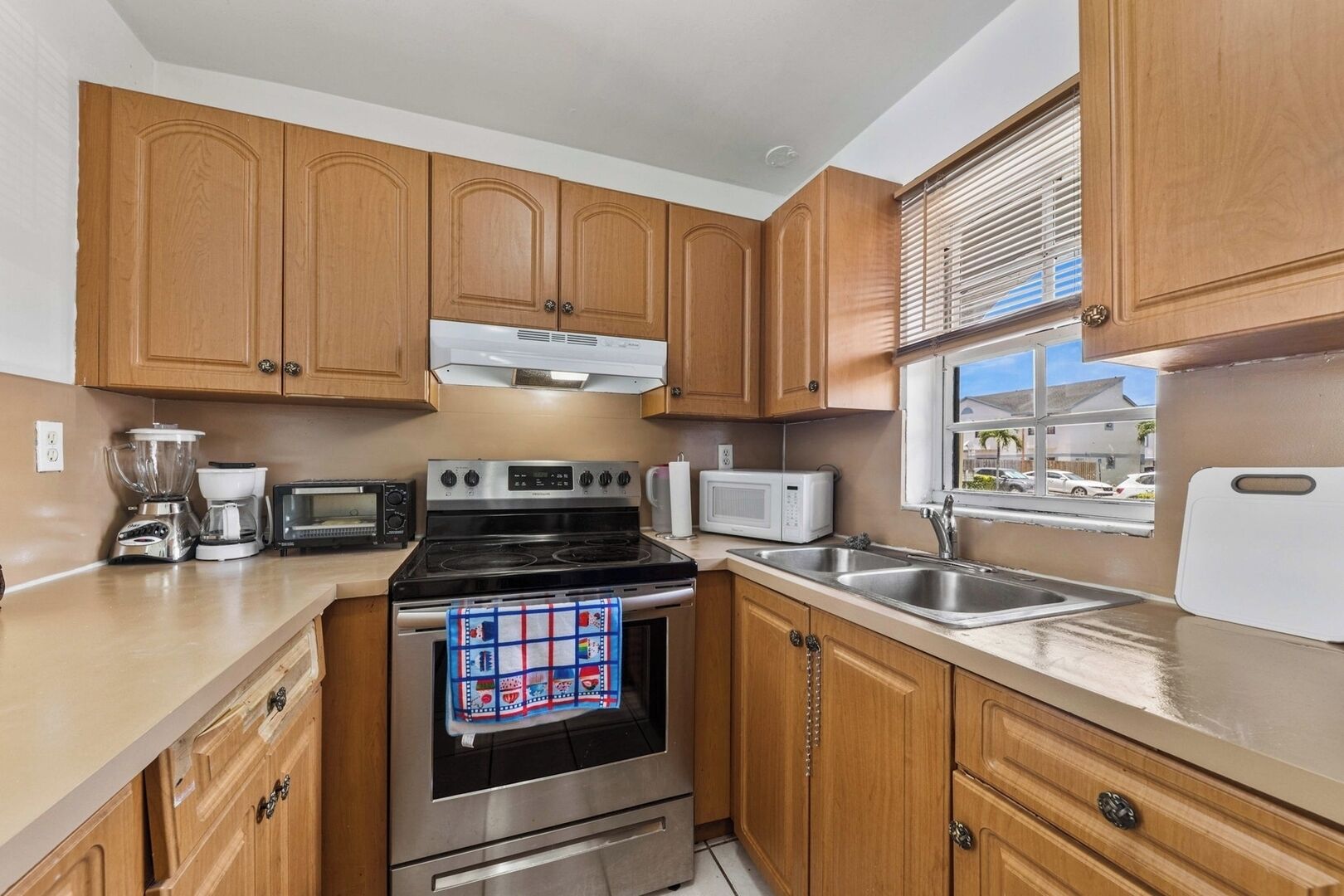 A compact kitchen with wooden cabinets, a stove, a microwave, a toaster, and a window with blinds.
