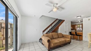 Un salón amplio con un gran ventanal, ventilador de techo, una escalera y una zona de cocina.