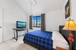 A bedroom with a bed, a desk with a television, a window with curtains, and a lamp.