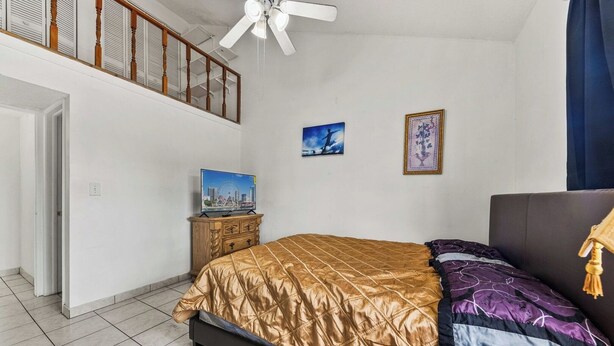 Un dormitorio con una cama, un televisor sobre un mueble de madera, un ventilador de techo y un balcón con barandas de madera.