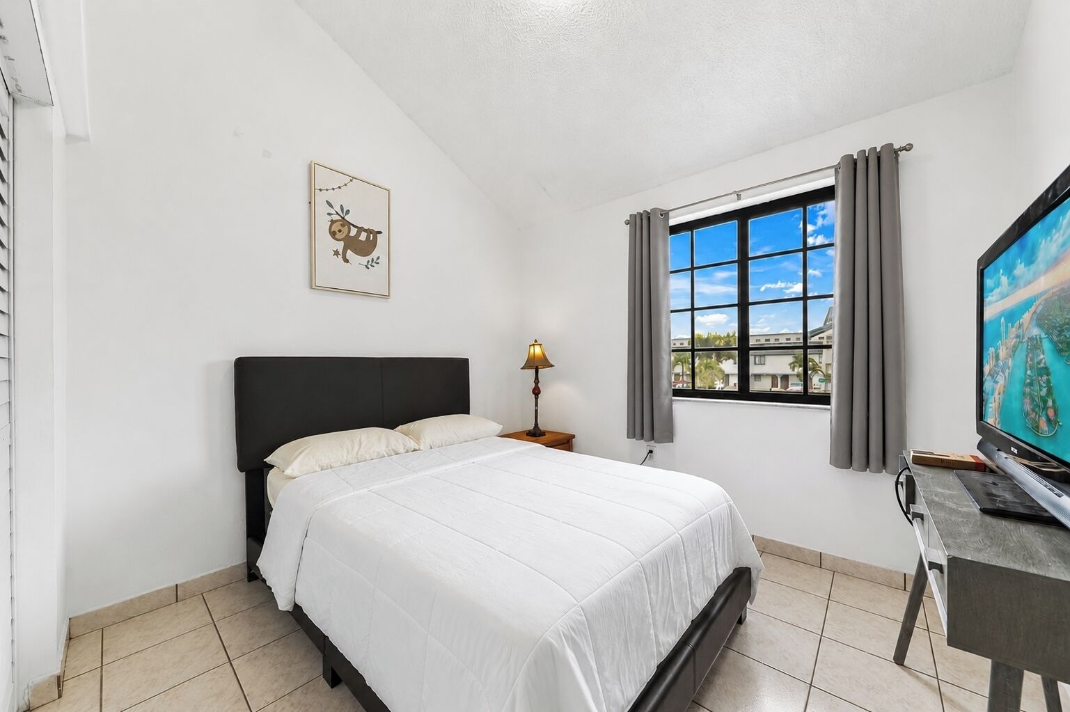 A hotel room with a bed, a desk with a television, a window with curtains, and a wall-mounted picture.