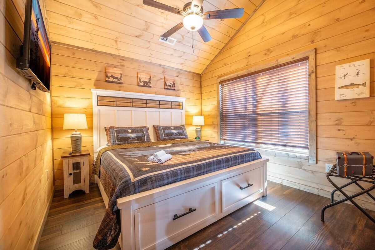Une chambre à coucher dans un chalet en bois, avec un lit, des lampes de chevet, un ventilateur de plafond et une fenêtre avec des stores.
