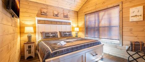 Une chambre à coucher dans un chalet en bois, avec un lit, des lampes de chevet, un ventilateur de plafond et une fenêtre avec des stores.