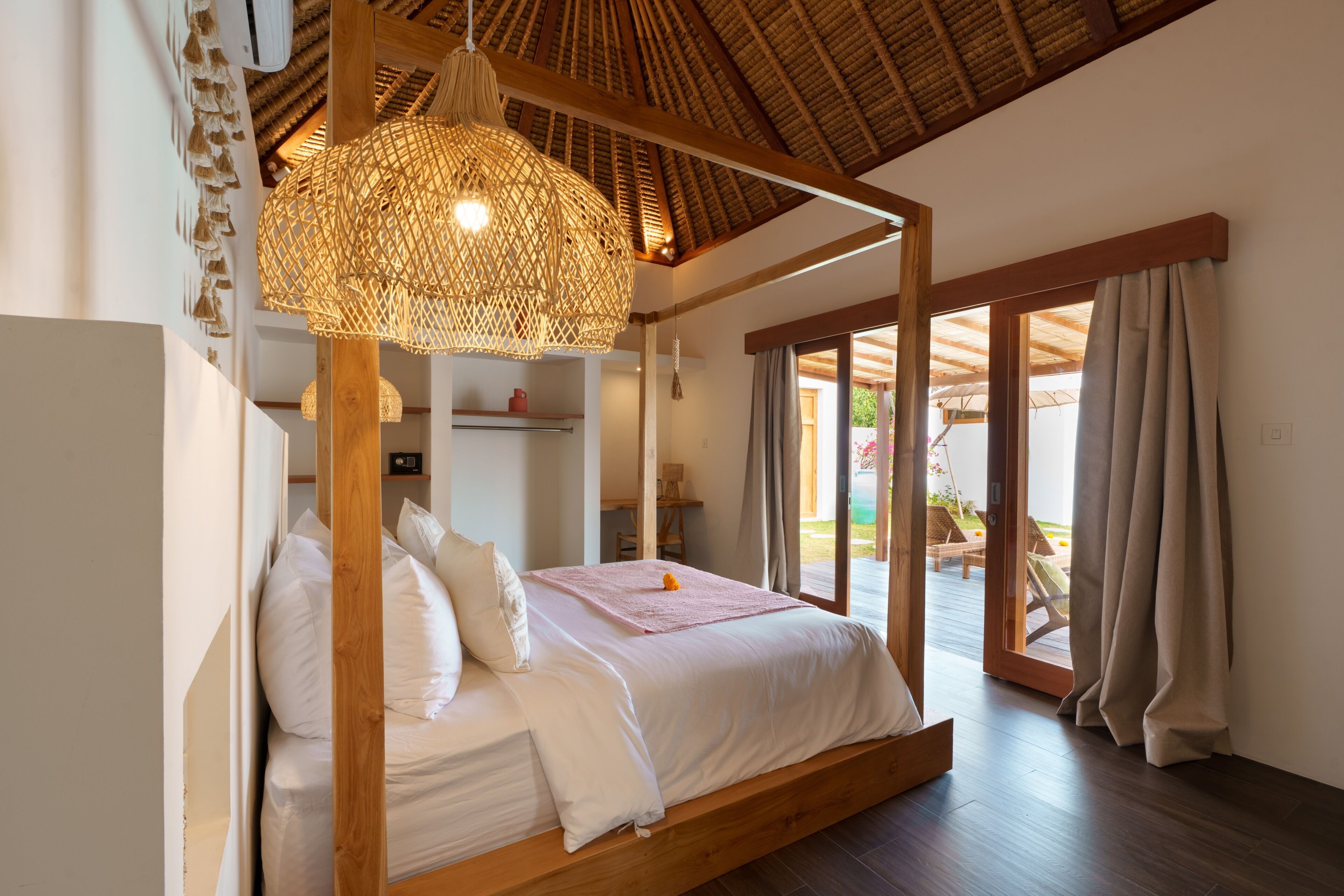 Een hemelbed met een geweven lamp boven, houten balken en uitzicht op een terras met lounge fauteuils.