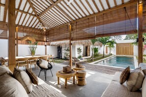 An indoor/outdoor living area with a pool, wooden furniture, and a thatched roof structure. - Villa Surya Tanah by Alfred in Bali (Seminyak)
