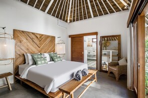 A modern bedroom with a wooden headboard, a large bed with white linens, a bench with towels, and a wicker chair. - Villa Surya Tanah by Alfred in Bali (Seminyak)