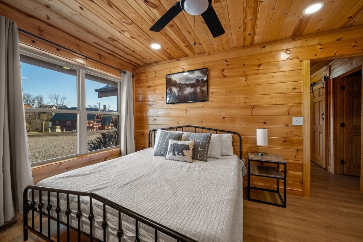 Une chambre à coucher aux murs lambrissés, avec un lit, un ventilateur de plafond, une fenêtre et un tableau accroché au mur.