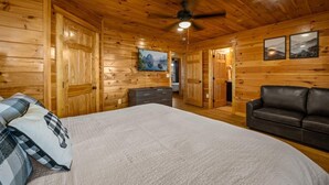 Une chambre dans une cabane en rondins, avec un lit, une commode, un canapé en cuir noir et un ventilateur de plafond.