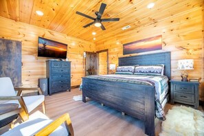 Ein Schlafzimmer in einer Blockhütte mit einem Bett, einem Schreibtisch, einem Sessel, einem Fernseher und einem Deckenventilator.