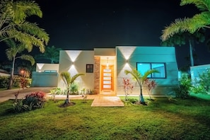 A modern house with a well-lit entrance, a stone wall, and a small garden with various plants. - Villa Tranquila, Luxury Boho Encuentro Retreat (sosua)