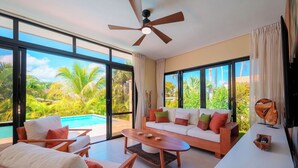 Ein geräumiger Wohnbereich mit großem Fenster, Deckenventilator, weißes Sofa mit bunten Kissen, Couchtisch aus Holz und ein Paar Holzschläppchen auf dem Boden.