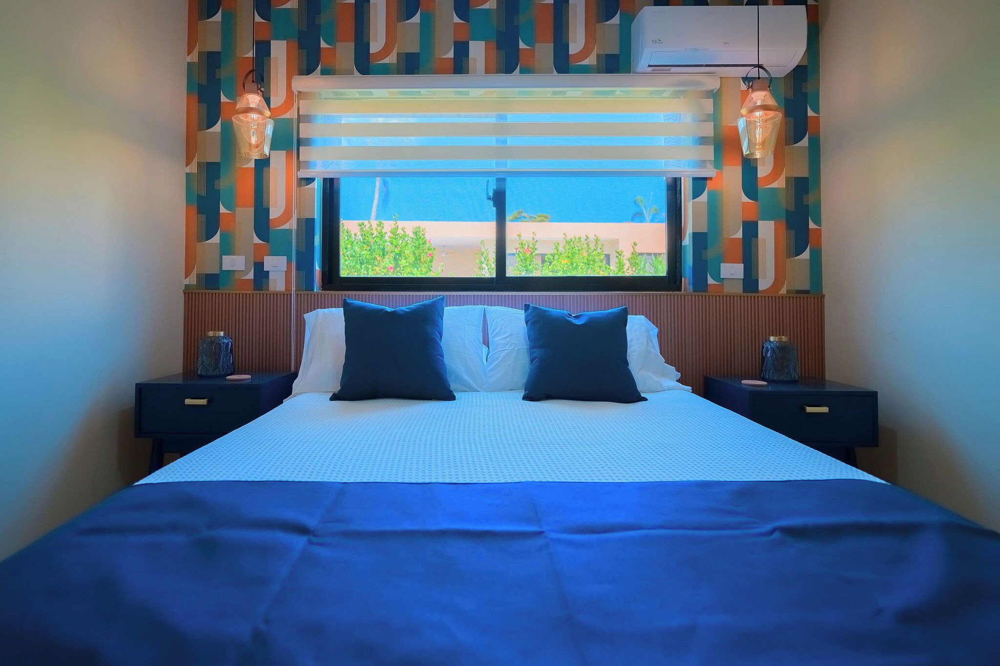Un letto rifatto con cura, con copriletto blu e due cuscini scuri, una finestra vista verde e una parete decorata.