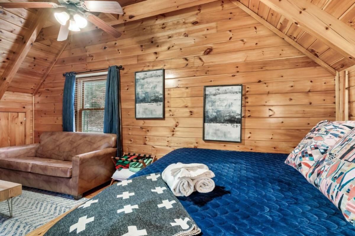 A cozy room with wooden walls, a bed with a blue quilt, a brown leather sofa, and a ceiling fan.