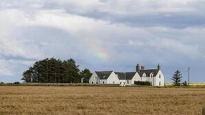 Un edifici gran i blanc amb diverses seccions, envoltat per un camp i arbres, sota un cel nuvolós.