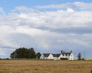 A large white building with multiple sections, surrounded by a field and trees, under a cloudy sky.