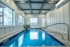 Uma piscina interior com água cristalina, uma escada de metal e vista para o exterior através de grandes janelas.