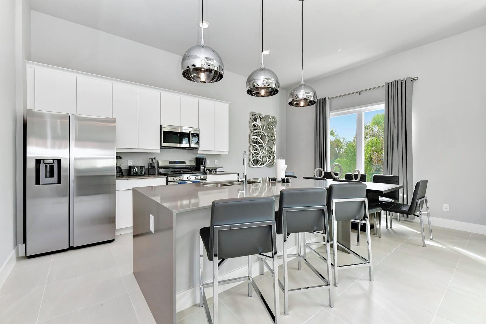A modern kitchen with stainless steel appliances, a central island with bar stools, pendant lighting, and a view of greenery outside.