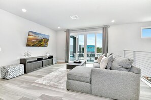 Ein modernes Wohnzimmer mit grauem Sofa, Couchtisch, Wandfernseher und großem Fenster mit Vorhängen.