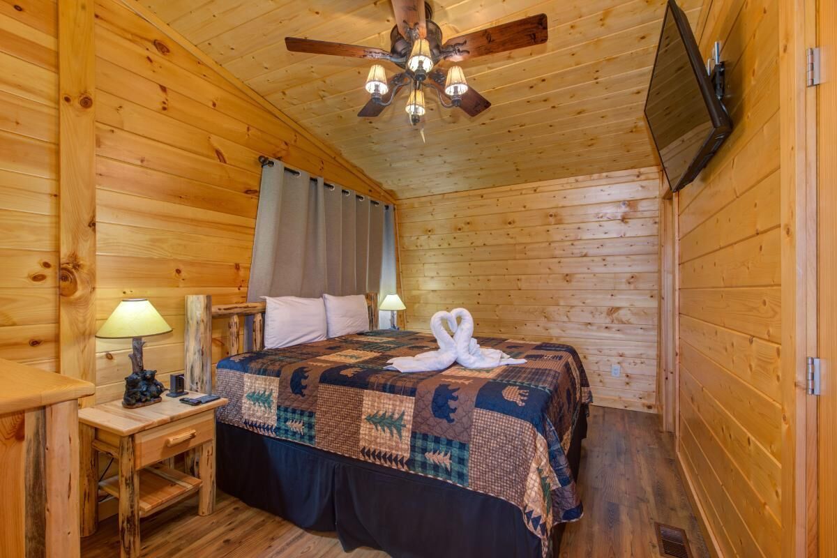 A wooden cabin room with a bed, two bedside lamps, a ceiling fan, and a flat-screen TV mounted on the wall.