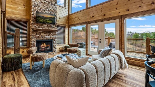 A spacious living room with a stone fireplace, wooden beams, and large windows overlooking a scenic landscape. - Arunachala (Sevierville)