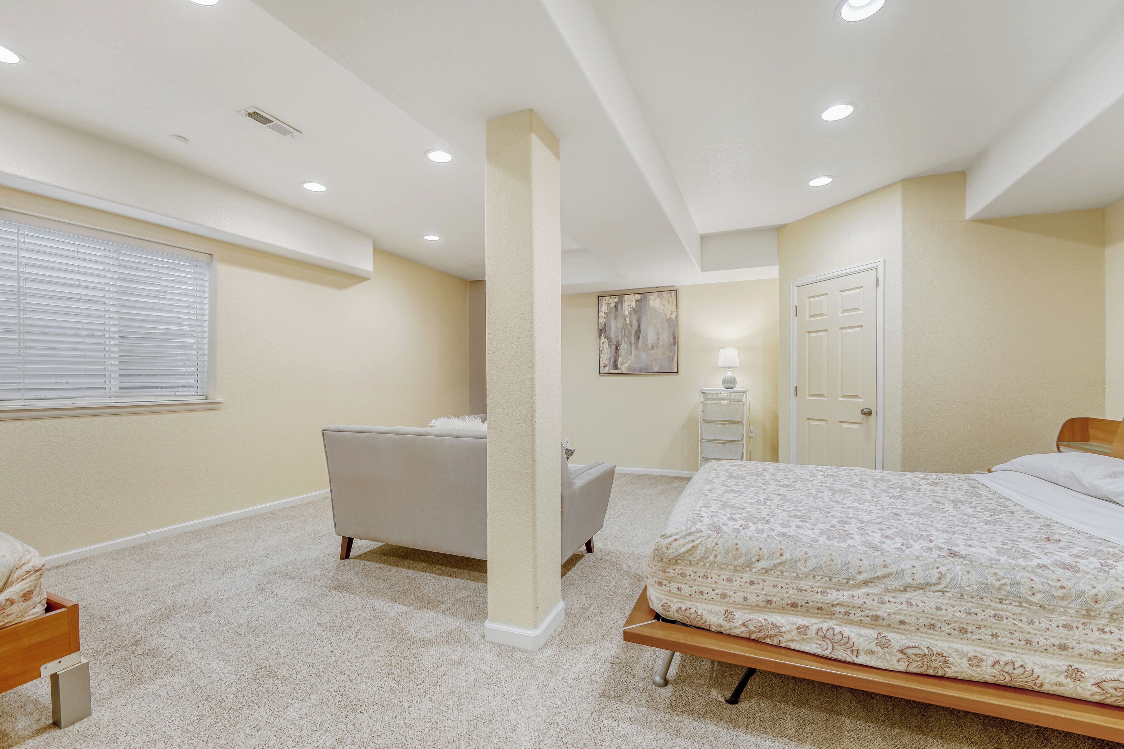 A bedroom with a bed, a vanity, a chair, a window with blinds, a door, and recessed ceiling lighting.