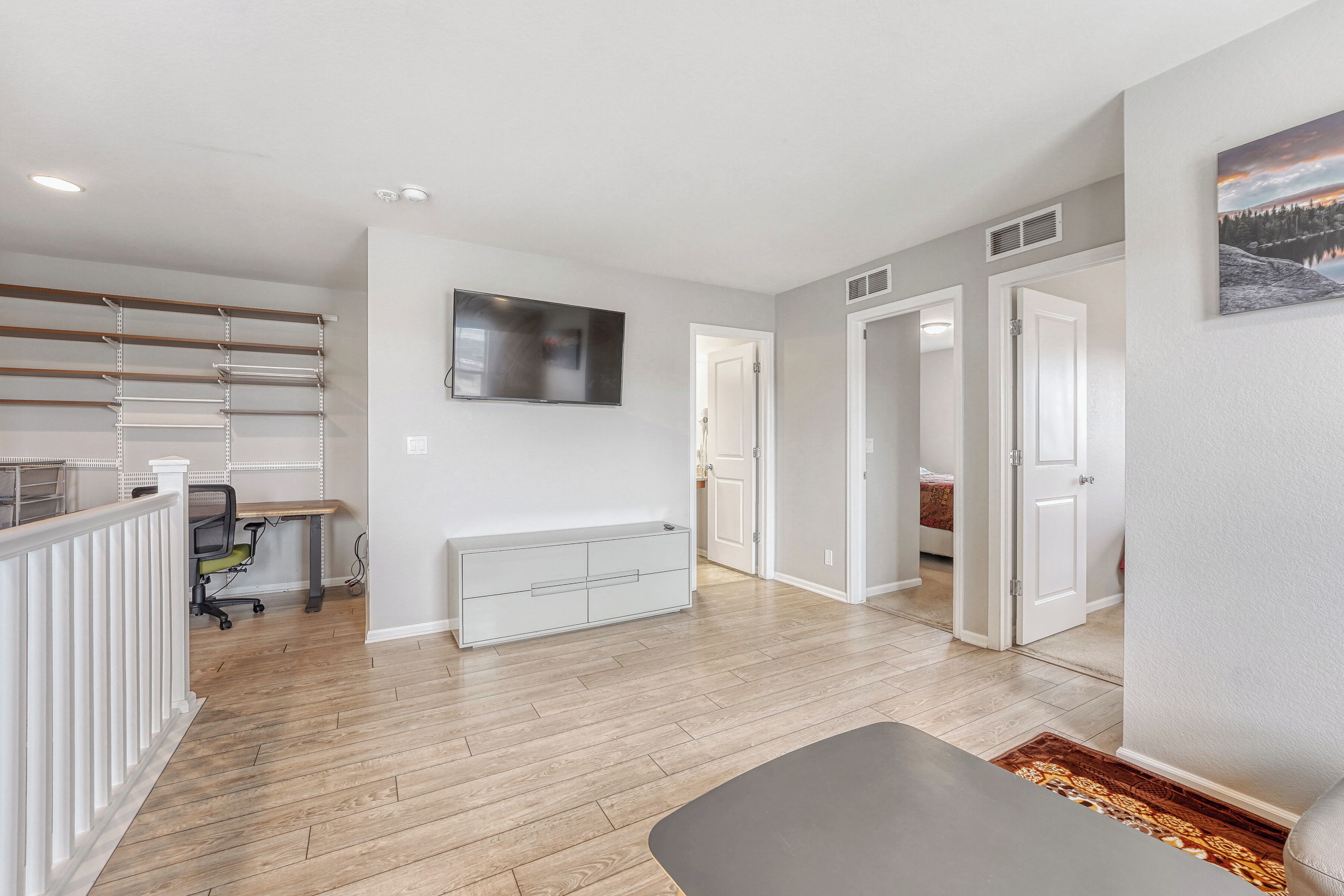 A spacious living area with a wooden floor, a grey sofa, a TV mounted on the wall, and a door leading to another room.