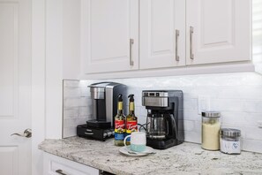 A kitchen with white cabinets, a coffee maker, two bottles of Torani syrup, a mug, and a container of sugar. - Capecation! A Boater's Paradise, easy & quick out. (Cape Coral)