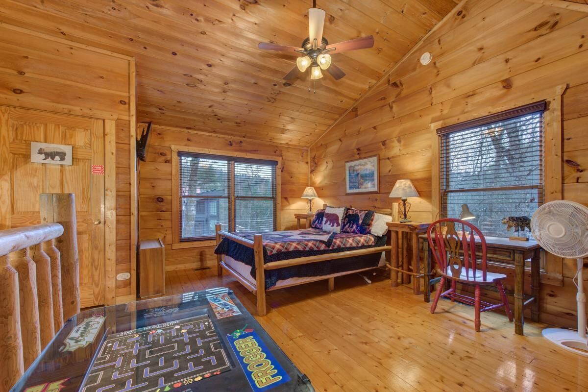 Una camera in una casetta di legno con un letto, una scrivania, una sedia, un ventilatore e una finestra.