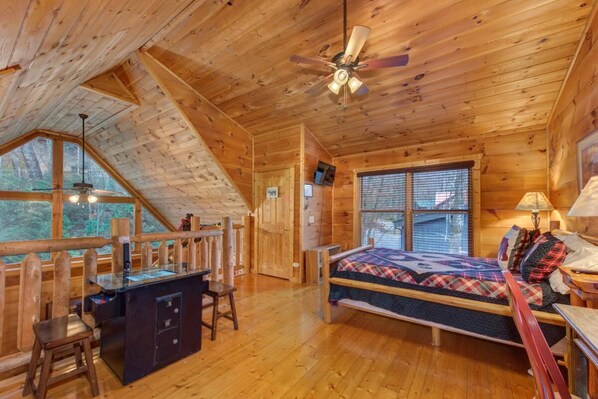 A log cabin bedroom with a bed, desk, and chair, a ceiling fan, and large windows. - Sleeping Bear Hideaway (Sevierville)