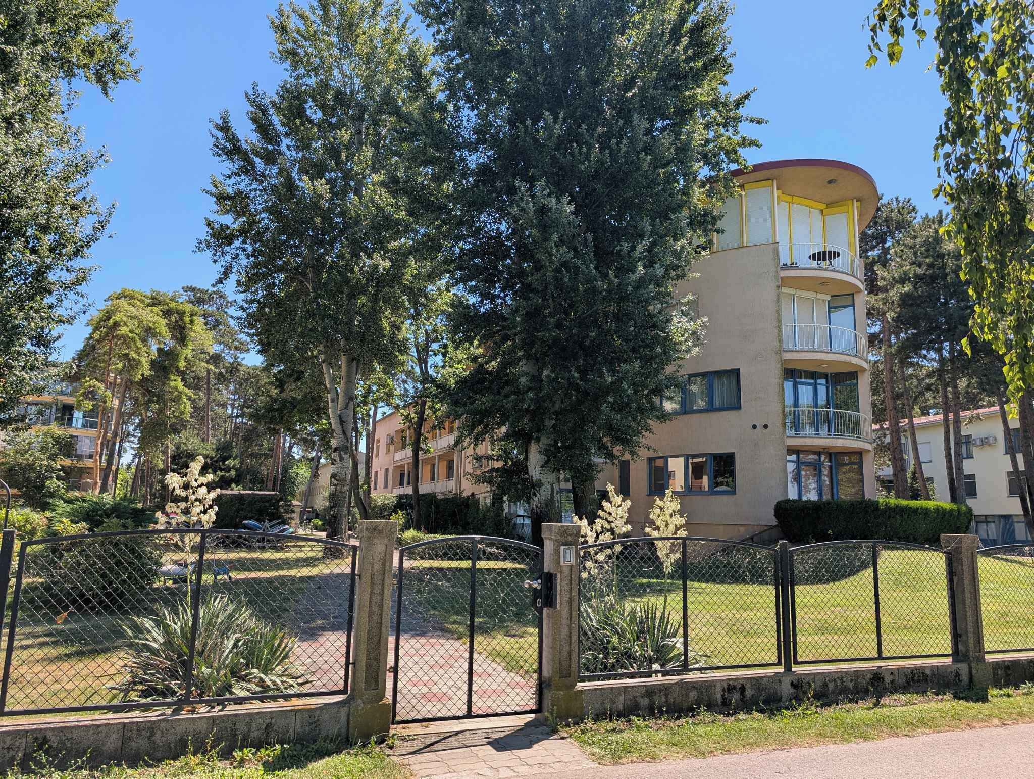 Un edificio de varios pisos con balcones, rodeado de árboles y un jardín cercado.