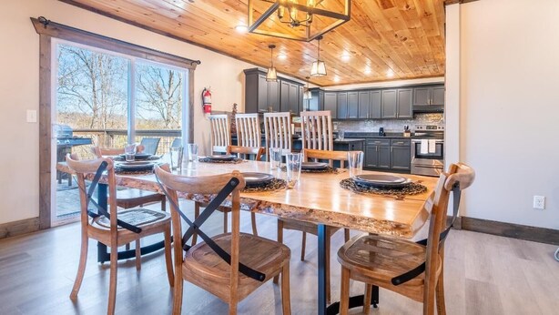 Une table à manger en bois, avec des chaises, un lustre et une vue sur l’extérieur.