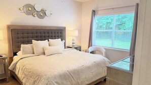 Un dormitorio con cabecera capitonada, una cama grande con ropa blanca, una mesita de noche con lámpara y una ventana con persianas.