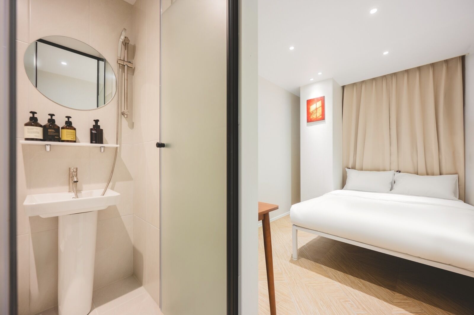A modern hotel room with a bathroom featuring a sink, mirror, and toiletries. The bedroom has a bed, curtains, and a small table.