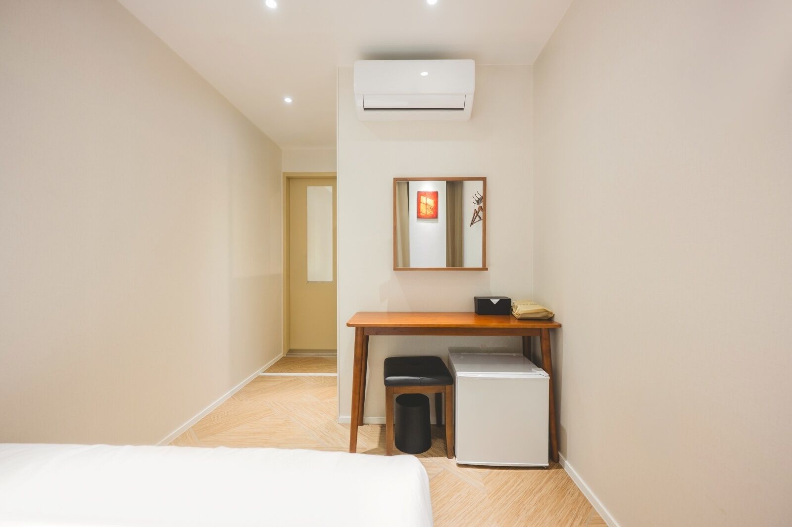A hotel room with a bed, a desk with a chair, a small refrigerator, and an air conditioning unit.