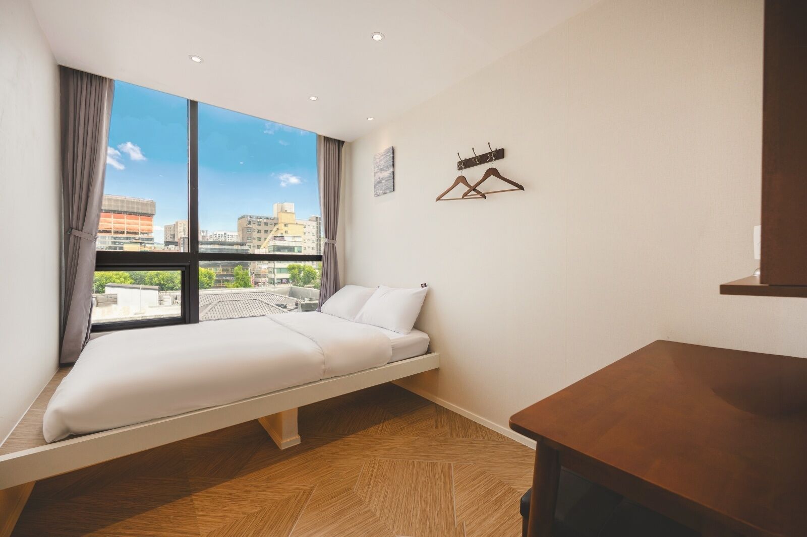 A modern hotel room with a bed, a small table, a view of the city, and a coat rack.