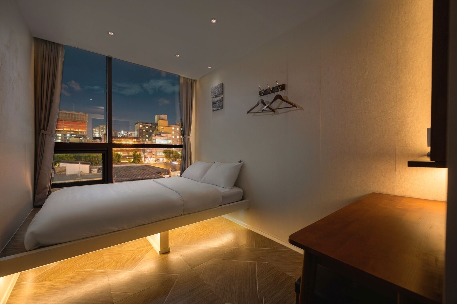 A modern hotel room with a bed, a small table, a view of the cityscape, and a wall-mounted coat rack.