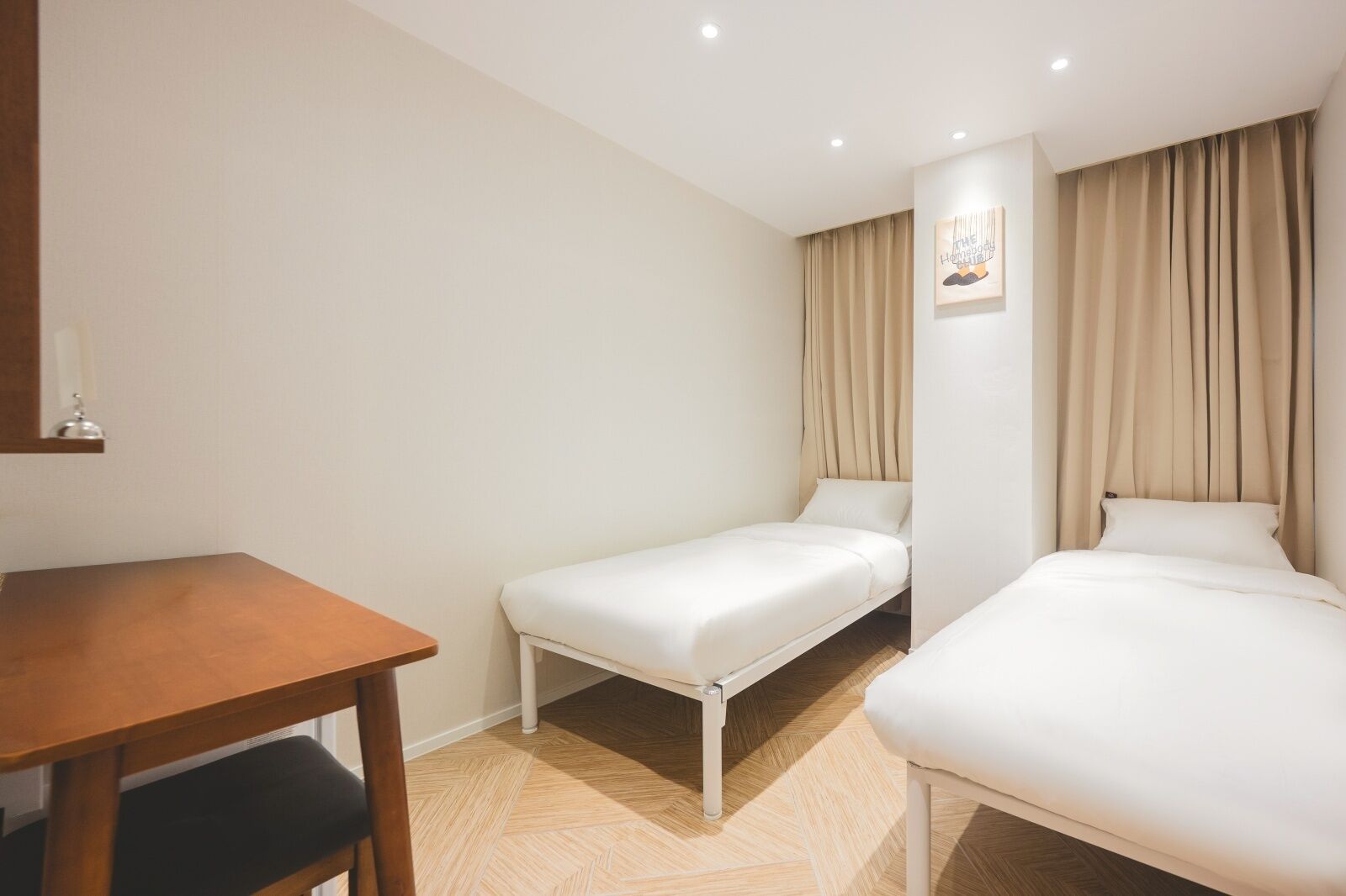 A hotel room with a wooden desk, two single beds, a small table, and a wall-mounted artwork.