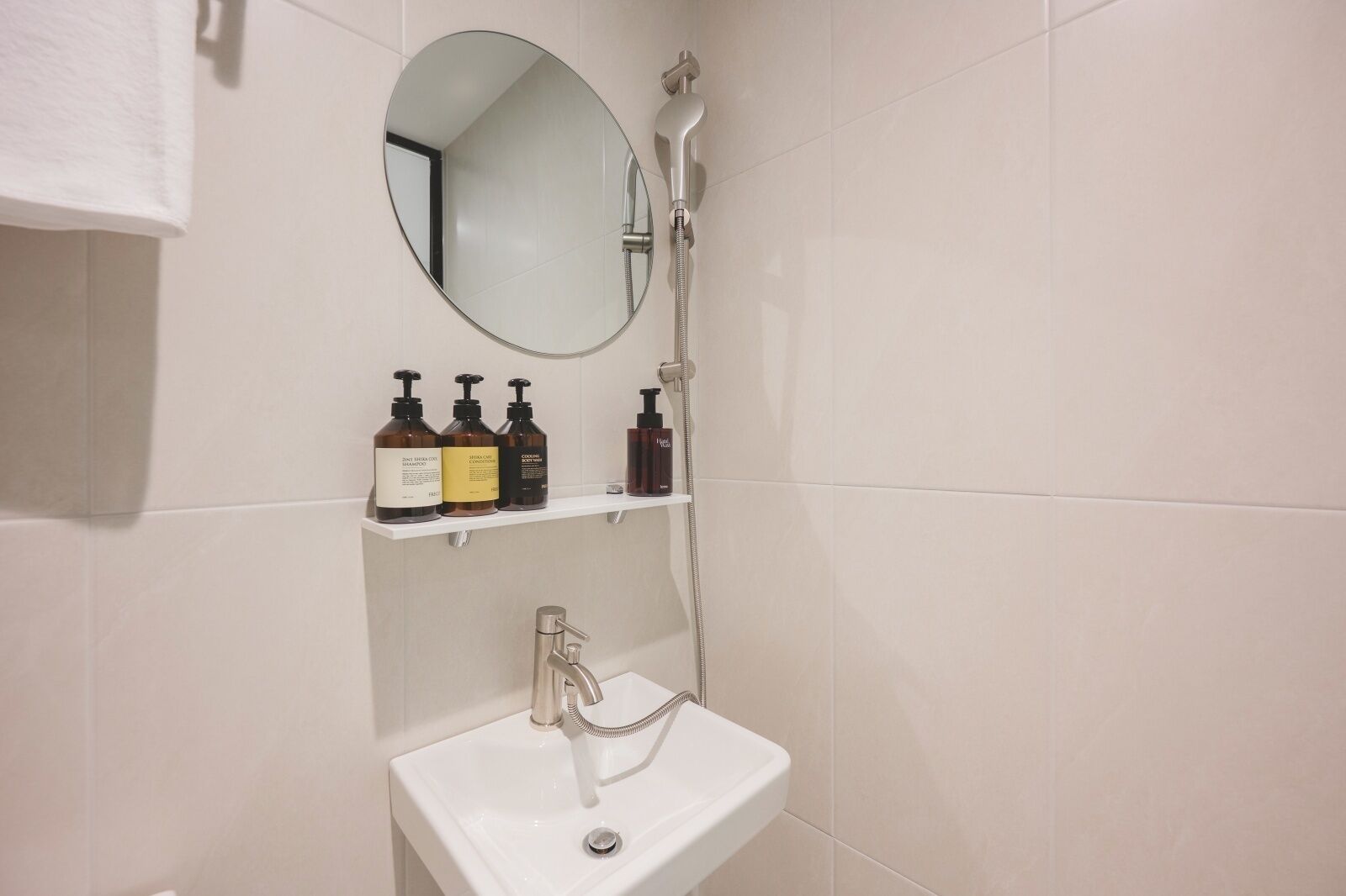 A bathroom with a white sink, a round mirror, and a shower. There are three bottles of shampoo on a shelf above the sink.