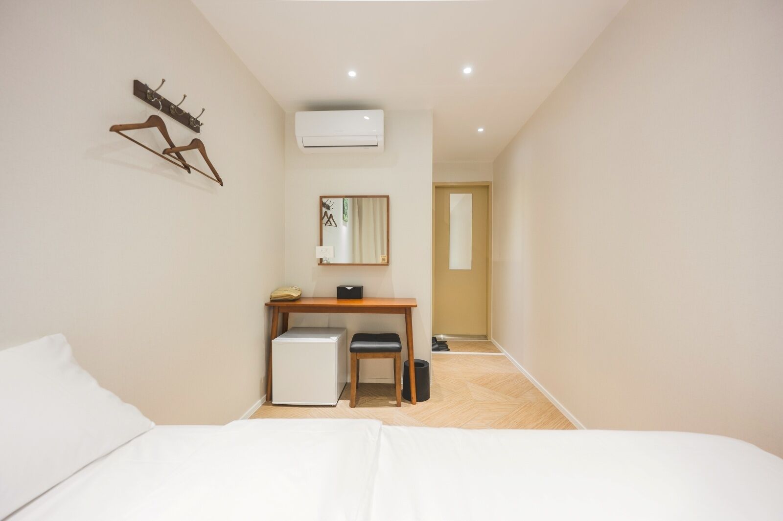 A hotel room with a bed, a desk with a chair, a small refrigerator, an air conditioner, and a coat rack.