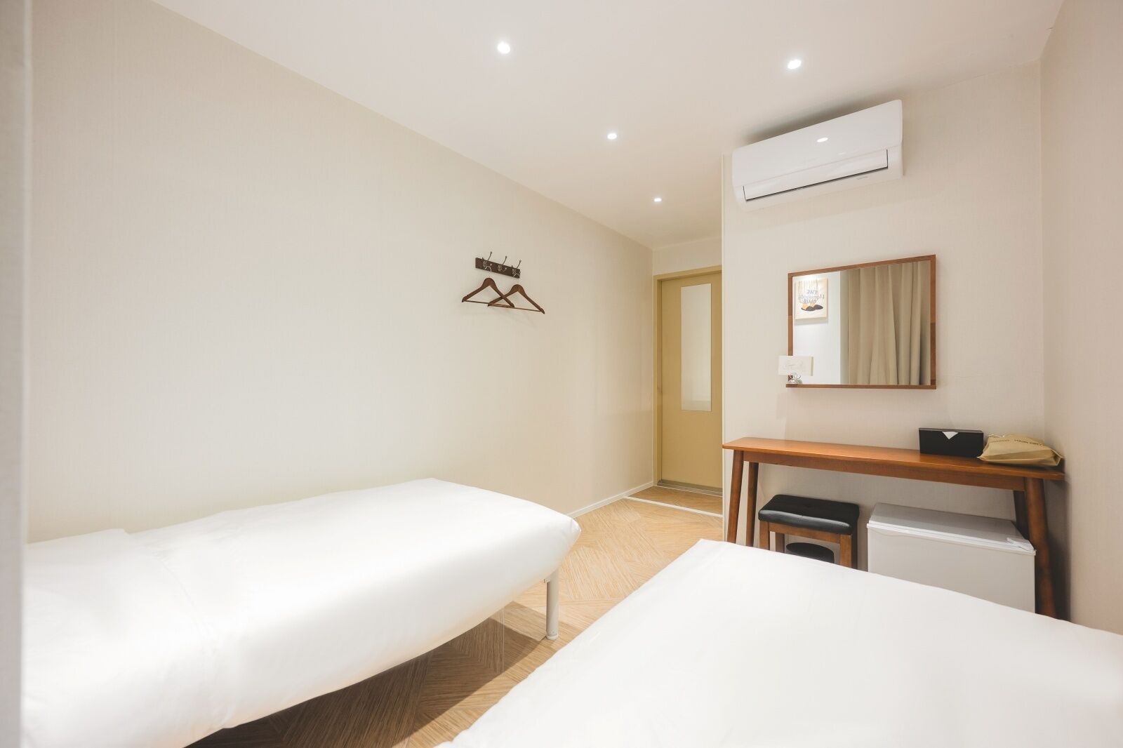 A hotel room with two single beds, a wooden desk, a small trash bin, an air conditioning unit, and a mirror.