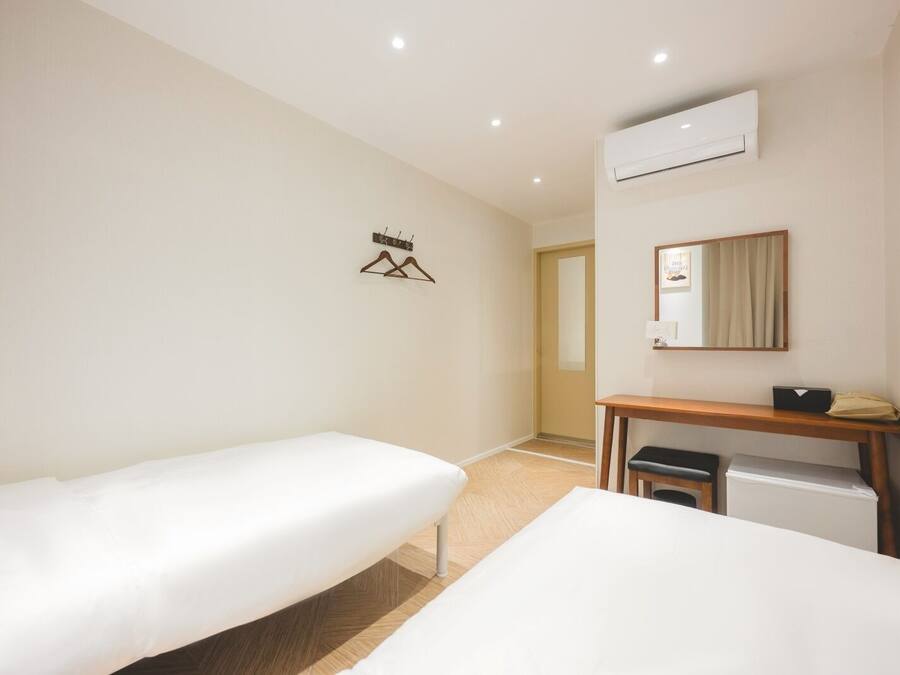 A hotel room with two single beds, a wooden desk, a small trash bin, an air conditioning unit, and a mirror.