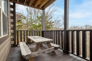 Eine Holzterrasse mit Picknicktisch, Geländer und Blick auf Bäume und ein Gebäude im Hintergrund.