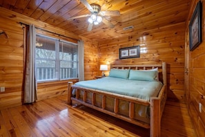Ein Schlafzimmer einer Holzhütte mit Bett, Deckenventilator, Fenster mit Jalousien und gerahmten Bildern an der Wand.