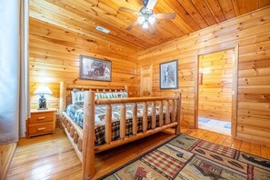 Een slaapkamer in een houten chalet, met een bed, een nachtkastje, een lamp, een plafondventilator en twee ingelijste foto's aan de muur.