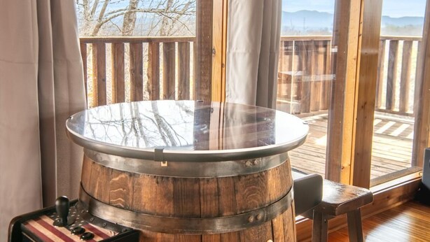 Ein Tisch aus Holzfässern mit Glasplatte, ein kleiner Hocker und ein Fenster mit Blick auf Bäume und eine Terrasse.