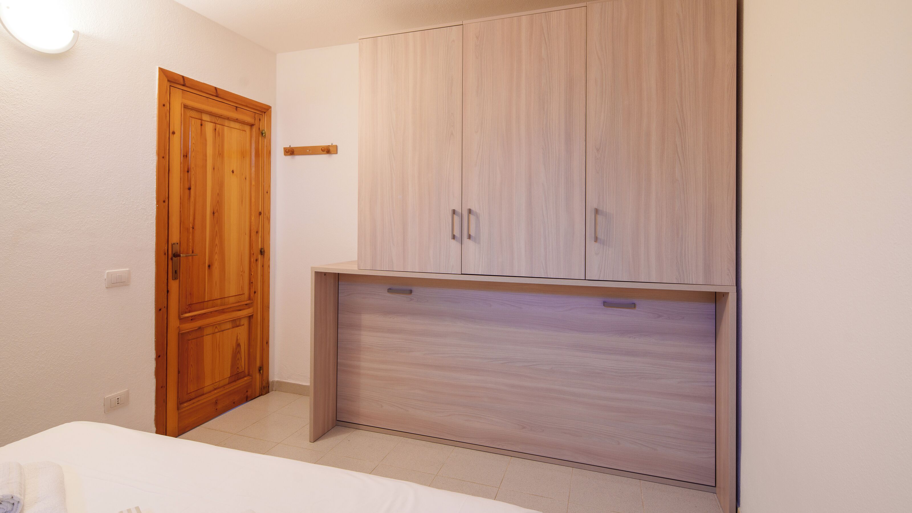 A room with a wooden wardrobe, a bed, and a light switch on the wall.
