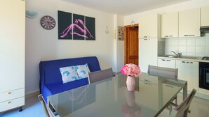 A modern living room with a glass dining table, a blue sofa with patterned cushions, a pink vase with flowers, and a kitchen in the background. - SpiaggiaSogno da Giò (San Teodoro)