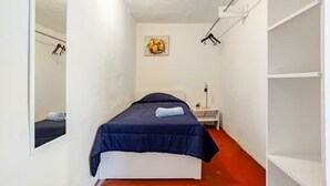 Ein schmales Zimmer mit einem Bett, einem Nachttisch, einem Spiegel und einem Wandbild.